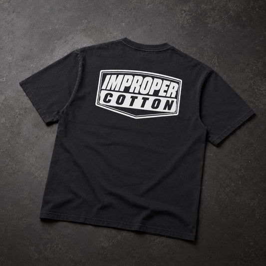 A black short-sleeve t-shirt laid flat on a dark textured background, featuring a white graphic print on the back that reads 'IMPROPER COTTON' inside a shield-like outline.
