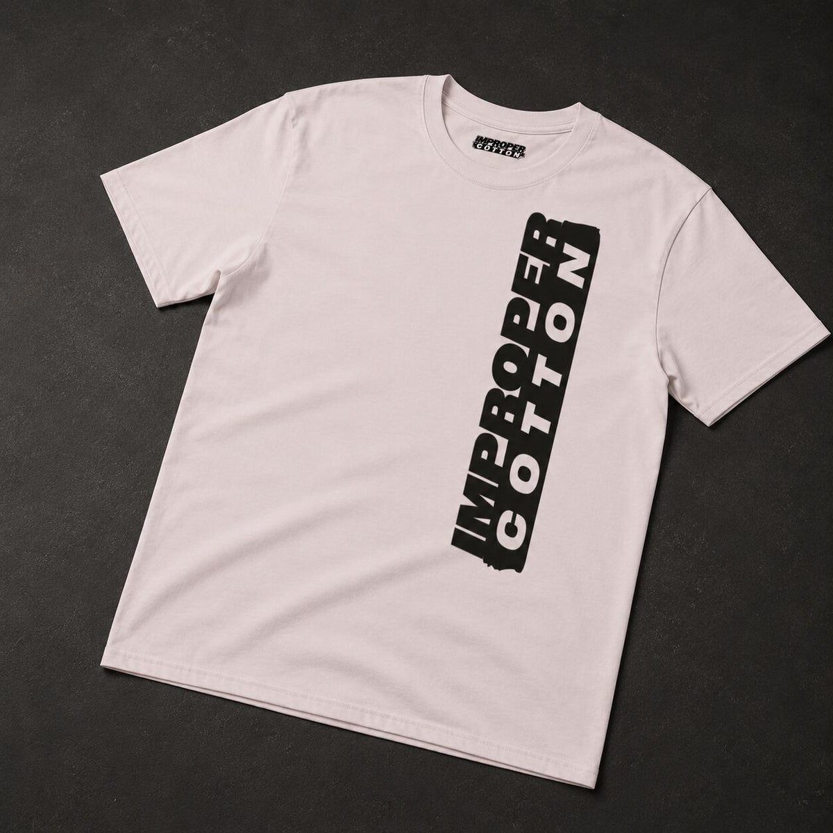 A light pink crewneck t-shirt with a vertical black graphic reading 'IMPROPER COTTON' on the front, laid flat on a dark textured background.