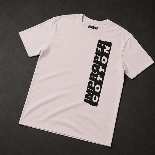 A light pink crewneck t-shirt with a vertical black graphic reading 'IMPROPER COTTON' on the front, laid flat on a dark textured background.