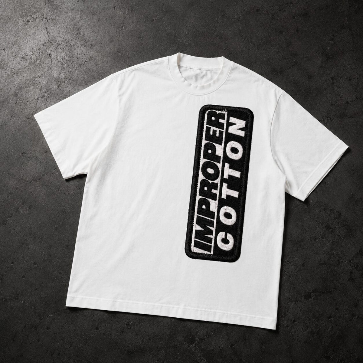 A white short-sleeve t-shirt with a vertical black graphic on the front reading 'IMPROPER COTTON' inside a rounded black rectangle, laid flat on a dark textured background.
