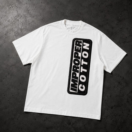 A white short-sleeve t-shirt with a vertical black graphic on the front reading 'IMPROPER COTTON' inside a rounded black rectangle, laid flat on a dark textured background.