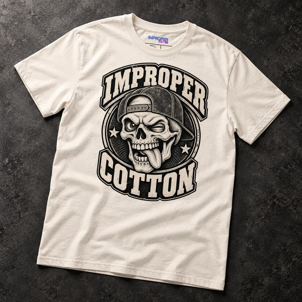 An off-white short-sleeve t-shirt with a black graphic print featuring a skull in a backwards cap and the text 'IMPROPER COTTON', laid flat on a dark textured background.