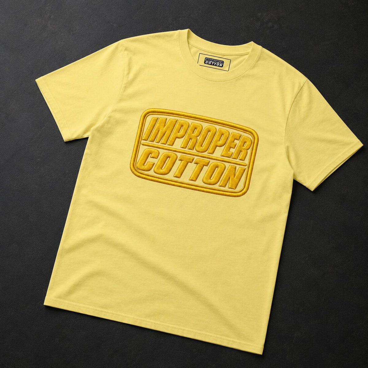 A pale yellow crewneck t-shirt with a large yellow and gold 'IMPROPER COTTON' graphic on the front, laid flat on a dark textured background.