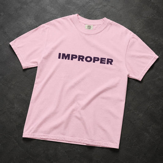 A light pink short-sleeve crewneck t-shirt with the word 'IMPROPER' printed across the chest in dark purple, laid flat on a dark textured background.