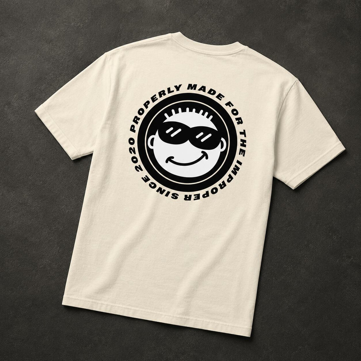 An off-white short-sleeve t-shirt laid flat on a dark textured background, showing a large black circular graphic on the back with a cartoon face wearing sunglasses and the text 'PROPERLY MADE FOR THE IMPROPER SINCE 2020'.