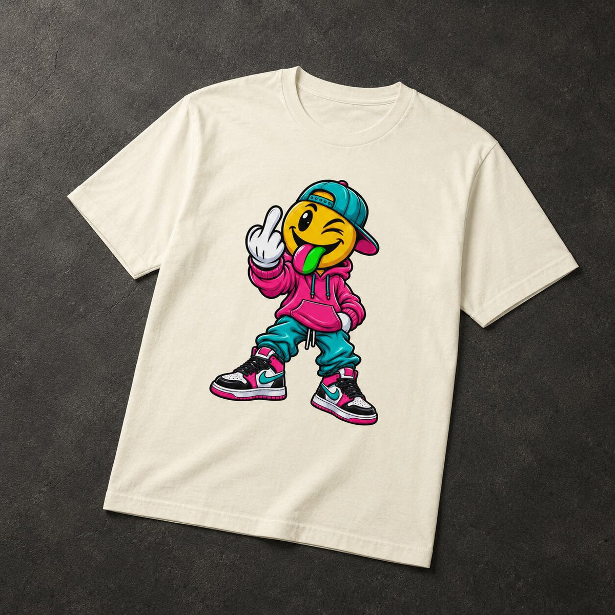 An off-white crewneck t-shirt laid flat on a dark textured background, featuring a colorful graphic of a cartoon emoji character wearing a pink hoodie, teal pants, and a blue cap.