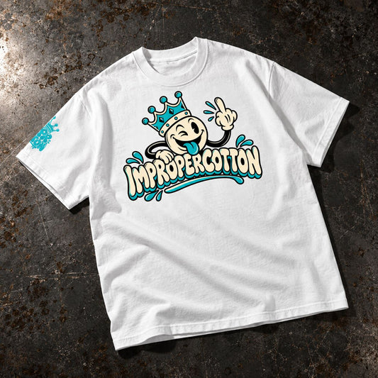 A white short-sleeve graphic T-shirt laid flat on a dark textured surface. The front features a cartoon character wearing a crown above the words 'IMPROPERCOTTON' in stylized lettering, with turquoise and cream details.