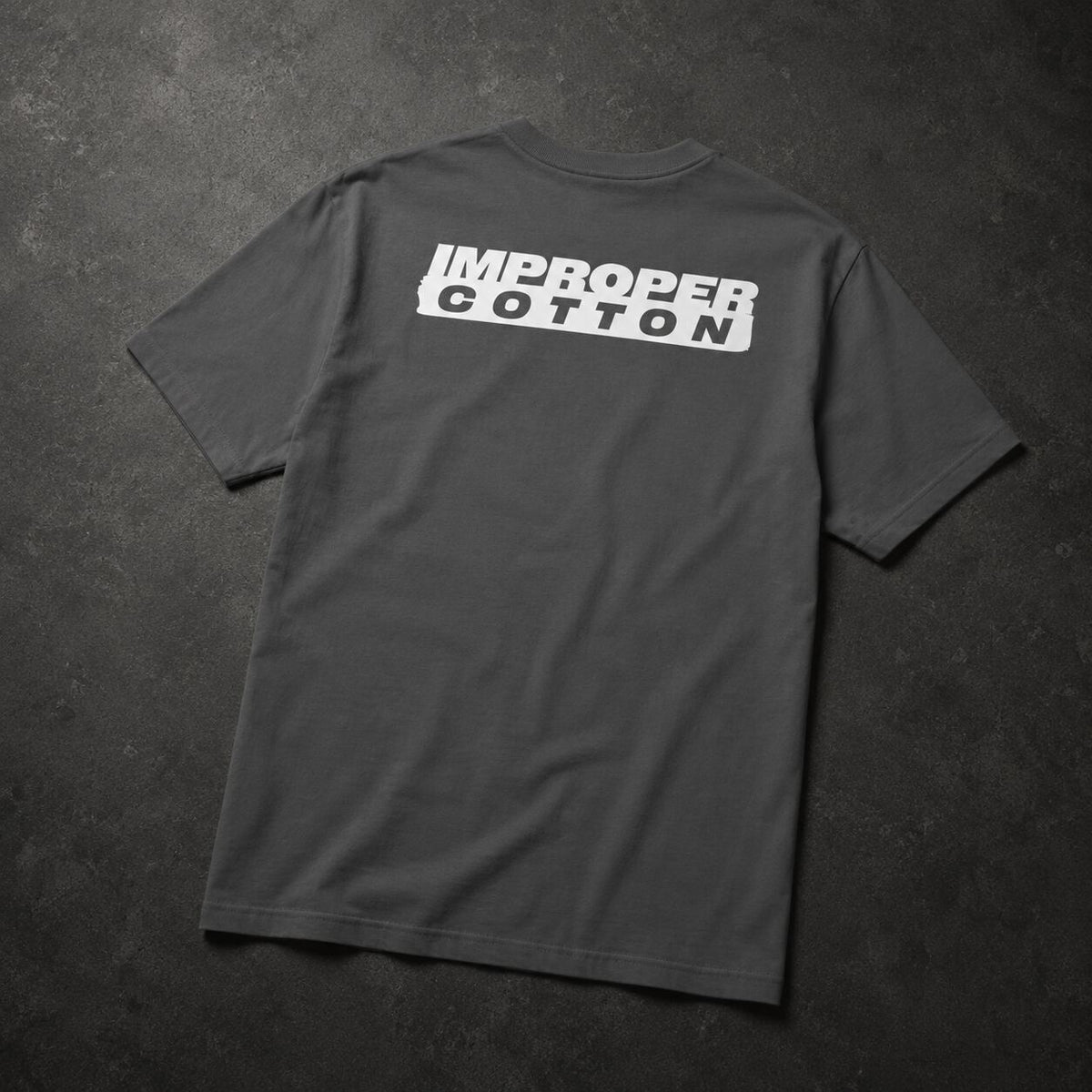 A dark gray crewneck t-shirt laid flat on a dark textured background, featuring a white graphic print on the back that reads 'IMPROPER COTTON' in a bold, stylized block font.