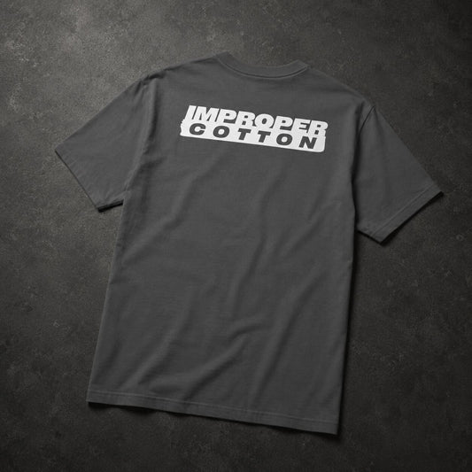 A dark gray crewneck t-shirt laid flat on a dark textured background, featuring a white graphic print on the back that reads 'IMPROPER COTTON' in a bold, stylized block font.