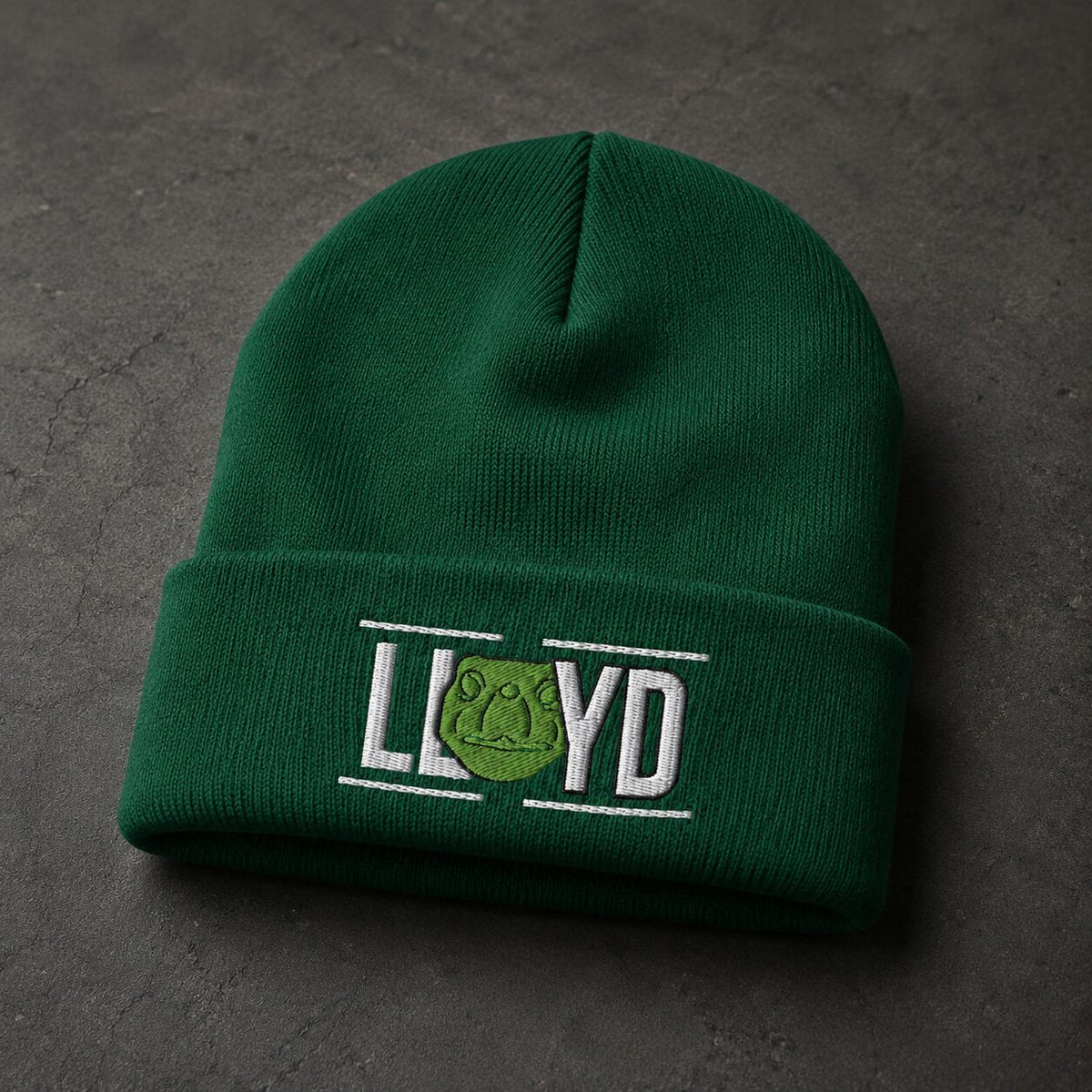 A dark green knit beanie hat with a folded cuff, featuring an embroidered logo that reads 'LLOYD' with a green frog emblem, placed on a dark textured surface.