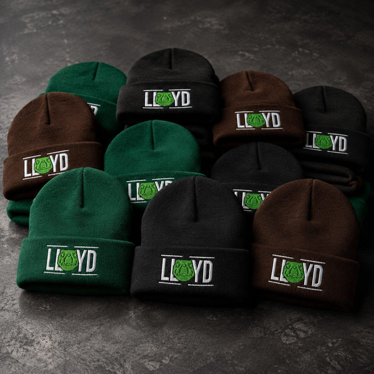 A group of knit beanie hats in green, brown, and black, each featuring a white embroidered 'LLOYD' logo with a green circular graphic on the cuff, arranged on a dark textured surface.