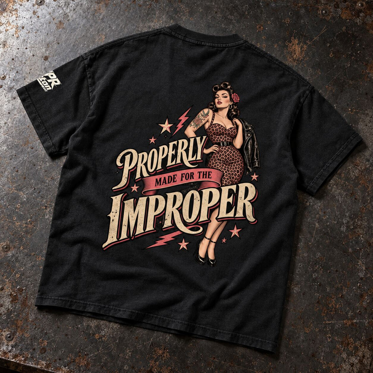 A black short-sleeve graphic t-shirt laid flat on a textured, rusty dark metal surface. The back of the shirt features a large retro-style pin-up illustration of a woman in a leopard-print dress with the text 'PROPERLY MADE FOR THE IMPROPER' in cream and pink lettering, accented with pink stars and lightning bolts.