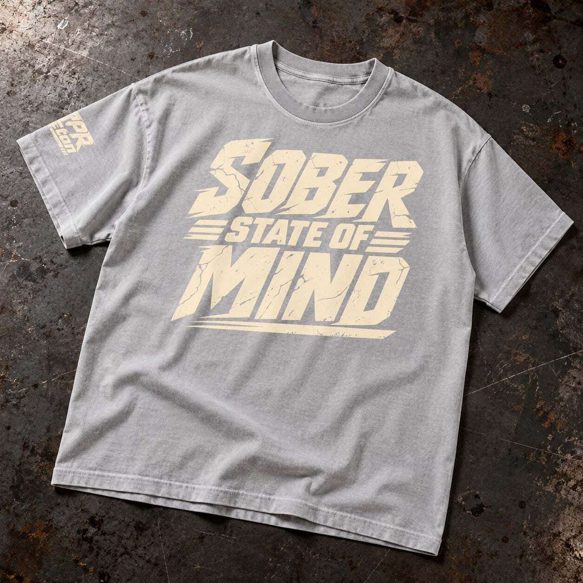 A light gray crewneck t-shirt with a distressed cream-colored graphic reading 'SOBER STATE OF MIND' across the chest, laid flat on a dark, textured industrial surface.