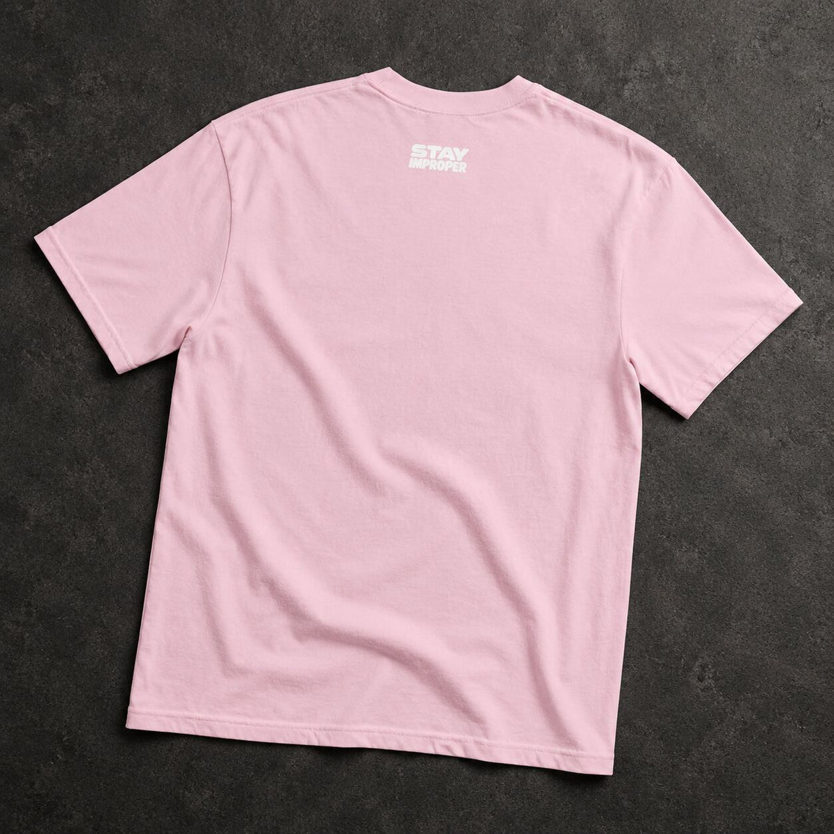 A light pink short-sleeve t-shirt laid flat on a dark textured background, featuring a small white graphic print on the upper back that reads 'STAY IN THE LOOP'.
